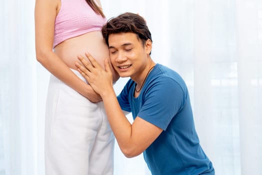 Asian man sit down and attach ear to belly of pregnant woman with smiling to express love and care in front of white curtain in their house.