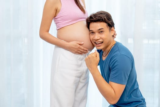 Asian man sit down and attach ear to belly of pregnant woman also look at camera show success emotion in front of white curtain in their house.