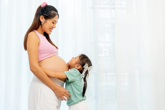 Asian little girl stand and hug belly of pregnant mother also look at her face in front of white curtain in their house.