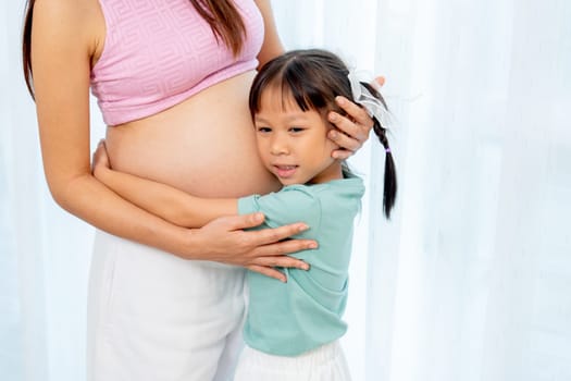 Asian little girl stand and hug belly of pregnant mother in front of white curtain in their house.