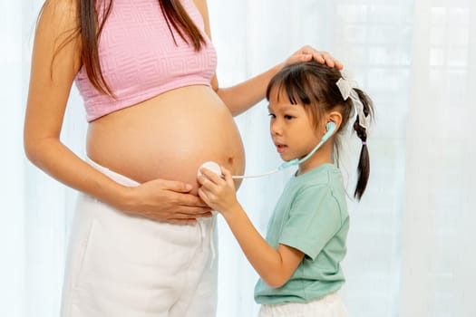 Close up Asian little girl use stethoscope toy to play with belly of pregnant mother in front of white curtain in their house.
