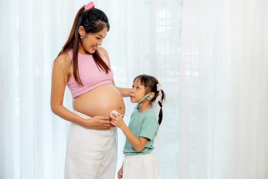 Asian little girl use stethoscope toy to play with belly of pregnant mother in front of white curtain in their house.