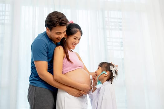 Asian father hug pregnant mother in her back and little girl use stethoscope toy to play with belly of mother in front of white curtain in their house.
