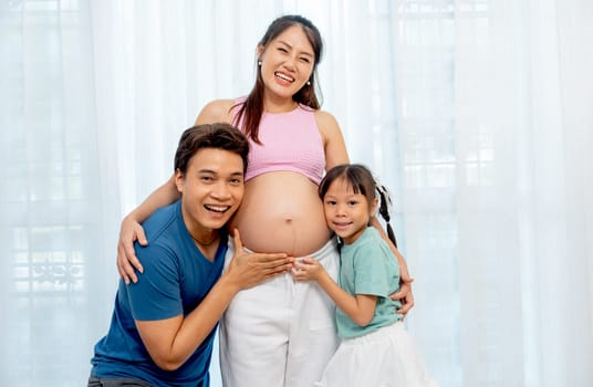 Asian father and little daughter hug and touch to belly of pregnant mother and express happiness with smiling in front of white curtain.