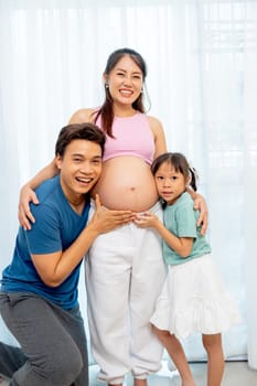 Vertical image of Asian father and little daughter hug and touch to belly of pregnant mother and express happiness with smiling in front of white curtain.