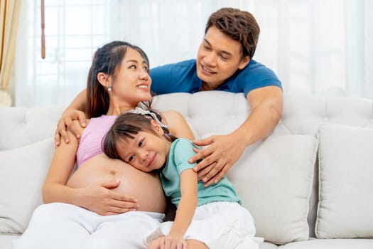 Main focus on Asian little girl lie and hug pregnant mother to listen sound and express care to little baby in belly of mother and father stay behind in concept of happiness family.