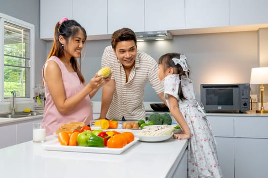 Asian pregnant mother show corn and play game to little girl and father stand beside and they look happy together in the kitchen.
