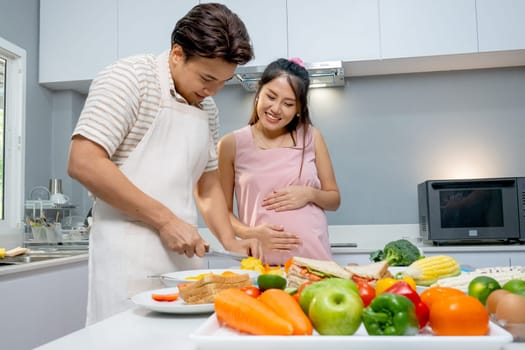 Asian man use knife to prepare or cook for pregnant woman who stand beside in the kitchen of their house.