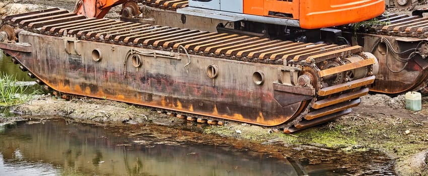 Closeup of a large, rusty crawler track, partially submerged in a muddy riverbank. The tracks are heavily weathered, showcasing their age and use.