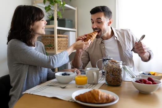 Happy couple having breakfast at home. Playful woman feeding croissant to husband. Lifestyle concept.