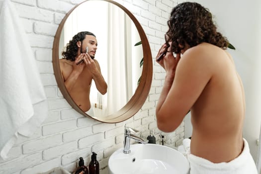 A man with curly hair grooming himself in the bathroom while looking into a round mirror close up