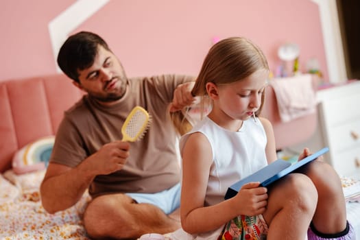 A man sits on a bed and brushes his daughter's hair. She is sitting beside him, reading a book. She is focused on the book while he brushes her hair.