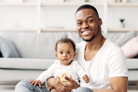 Babies Development. Portrait Of Happy Young Black Father With Little Baby On Lap Relaxing On Floor In Living Room, Cheerful African American Dad Playing With HIs Adorable Infant Child At Home