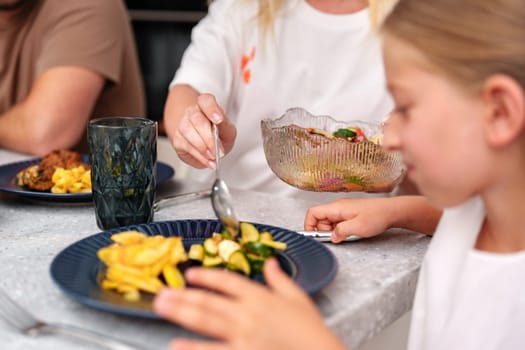 A girl with blond hair sits at a table and eats from a blue plate. Her hand rests on a blue and silver fork. In front of her is a plate with yellow potatoes and green vegetables.