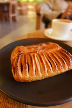 A freshly baked and flaky pastry presented on a dark plate next to a steaming cup of coffee