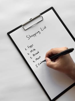 Hand writing shopping list on clipboard with eggs, milk, bread, meat, cakes items.