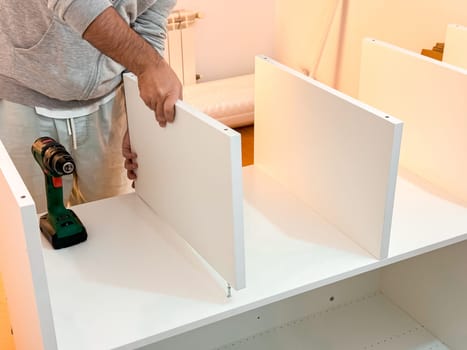 Caucasian male adult assembling white shelving unit with electric drill at home.