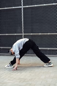 A person wearing a gray sweatshirt and black pants is stretching on pavement in front of a chain link fence. They are leaning forward, reaching their hands down to touch the ground.