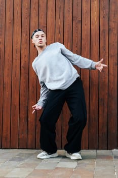 A young man wearing a bandana, gray sweatshirt, and black pants is dancing in front of a brown wooden wall. He is bending his knees and moving his arms in a fluid motion.