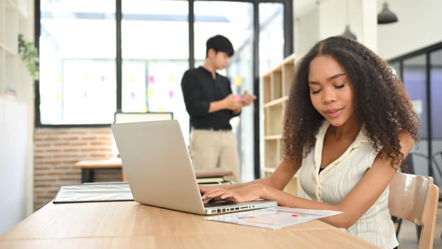 African American businesswoman reviewing charts and working on laptop at modern office desk.