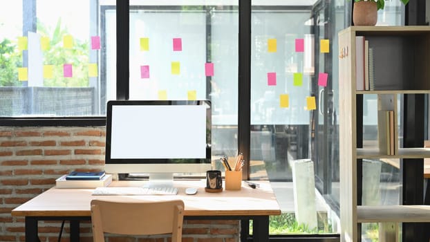 Bright office desk with blank desktop computer and stationery against brick wall and windows filled with sticky notes.