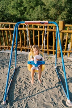 Little smiling girl swinging on a swing hugging a pink bunny at the playground. High quality photo