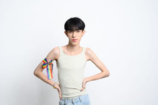 Confident young gay man with rainbow ribbon tied on arm standing on white background. LGBTQ pride and support.