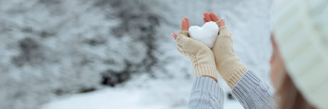 banner Close-up Snow heart in hands in knitted gloves on the background of snow cover. Valentine's Day.