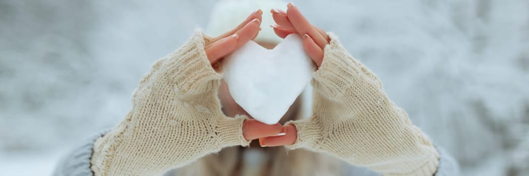 banner Close-up Snow heart in hands in knitted gloves on the background of snow cover. Valentine's Day.