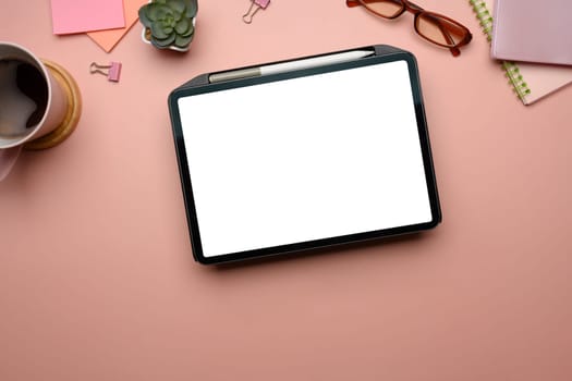 Feminine workspace with digital tablet, coffee cup, notebooks and accessories on pastel pink background.