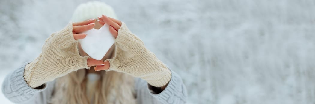 banner Close-up Snow heart in hands in knitted gloves on the background of snow cover. Valentine's Day.