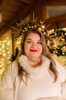 A woman wearing a white sweater and a gold crown with ornaments smiles at the camera. A Christmas wreath is visible in the background.