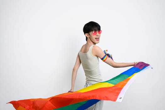 Joyful asian man wearing pink sunglasses swirling with pride flag against white background. LGBTQ pride and support.