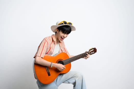 Joyful teenage man wearing hat and sunglasses playing guitar against white background.