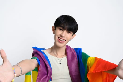 Cheerful man wrapped with pride flag standing over white background. LGBTQ equality and support concept.