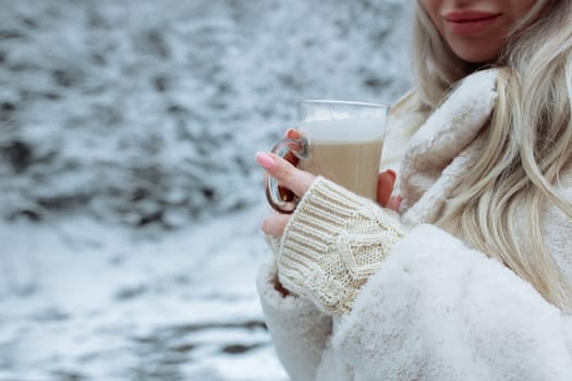 Beautiful hands hold a cup of cappuccino against the background of the winter forest. Gently beige gloves on the hands of a girl in the hands of coffee.