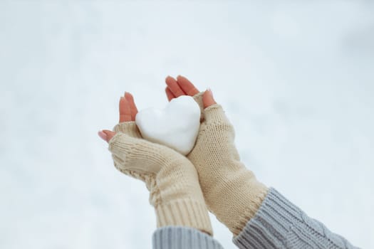 Close-up Snow heart in hands in knitted gloves on the background of snow cover. Valentine's Day.