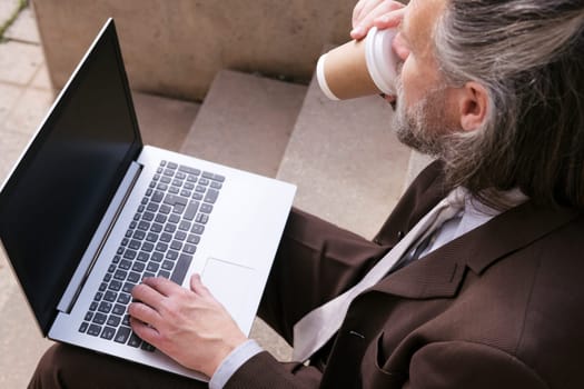 unrecognizable middle age male entrepreneur in formal suit sitting on urban stairway drinking takeaway coffee and working online on laptop in city, concept of business and mature man lifestyle