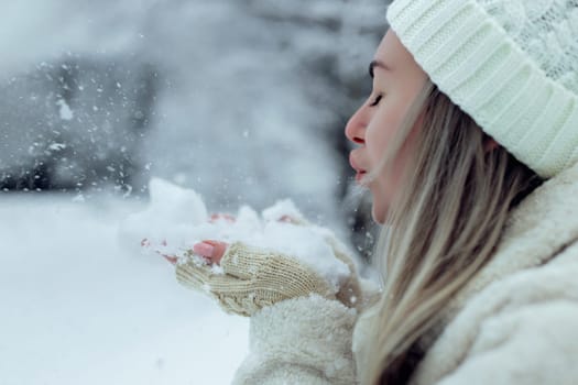 Beautiful girl in fashionable winter clothes blows snow from the palms of her hands. Joy to the first snow .Young blonde in a white hat in a snowy forest.