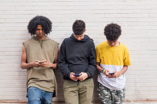 three serious diverse teens addicted to mobile phones browsing Internet while leaning on brick wall, concept of youth and technology of communication