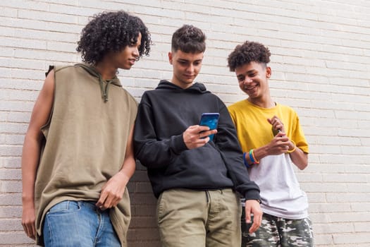 three smiling diverse teens having fun using mobile phone while browsing internet while leaning on brick wall, concept of youth and technology of communication