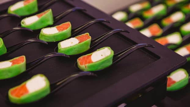 Platter of gourmet appetizers featuring pieces of salmon and cream cheese wrapped in a green wrap, served on black spoons. The presentation is elegant and suitable for catering events or parties