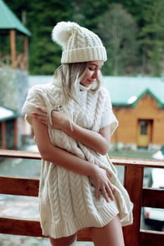 A sexy girl in a white sweater and stockings on the balcony admires the beauty of nature. wool clothes and a hat on a beautiful blonde.