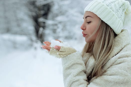 Beautiful girl in fashionable winter clothes blows snow from the palms of her hands. Joy to the first snow .Young blonde in a white hat in a snowy forest.