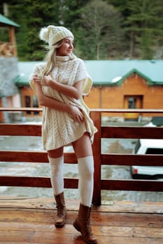 A sexy girl in a white sweater and stockings on the balcony admires the beauty of nature. wool clothes and a hat on a beautiful blonde.