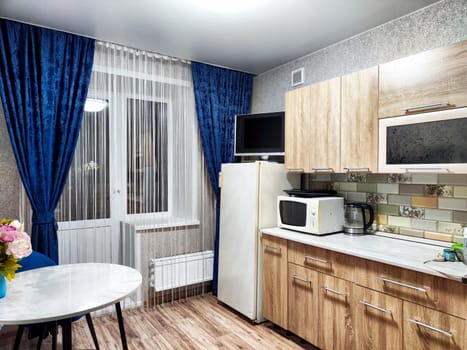 Cozy kitchen showcasing wooden cabinets, modern appliances, blue curtains, and a small dining table near the window.