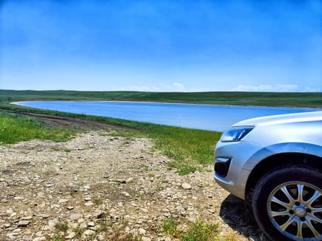 A silver car is parked near a tranquil lake surrounded by lush greenery under a clear blue sky on a bright day.
