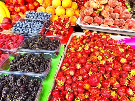 Brightly colored berries and fruits fill the market stand, showcasing strawberries, blueberries, and raspberries in midday light.