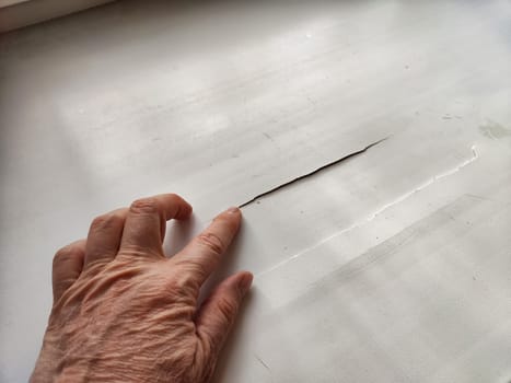 A hand gently touches a cracked window sill, highlighting the damage and wear in a home setting that's seen better days.