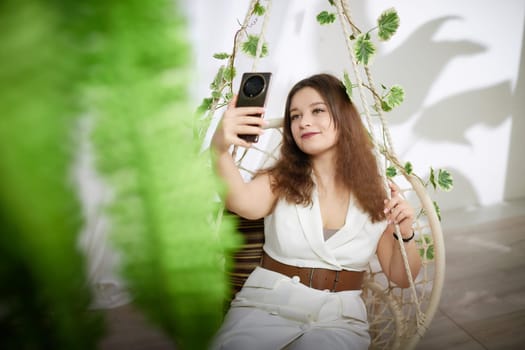 A woman sits in a hanging chair, enjoying her surroundings while capturing a selfie with her smartphone.